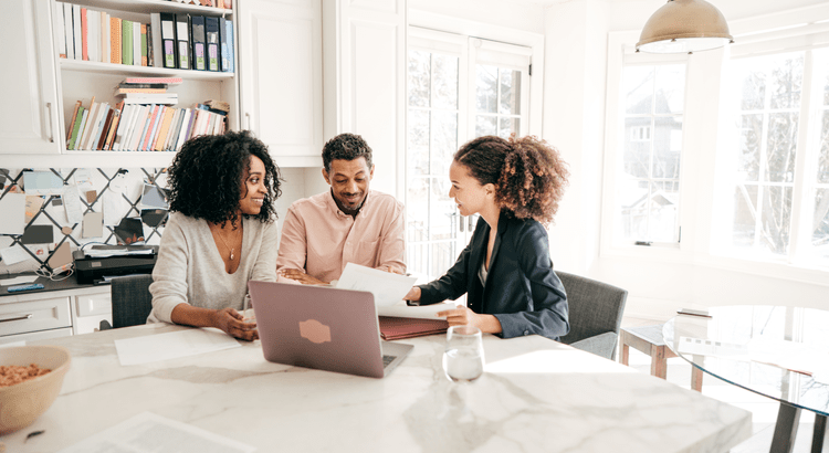 Two homeowners review documents with a real estate agent at a kitchen table.