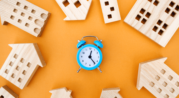 Wooden house models surround a blue alarm clock on an orange background.