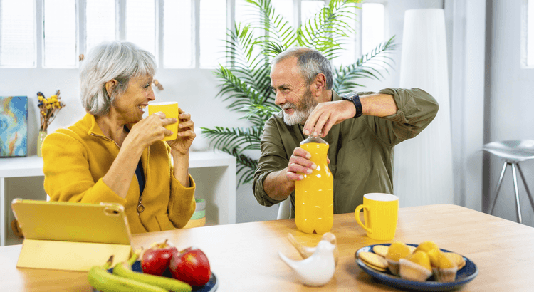 Older couple sharing breakfast at a kitchen table with yellow mugs and orange juice.