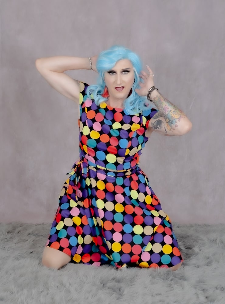 A drag queen with bold blue hair confidently poses in colorful, playful polka dot attire.