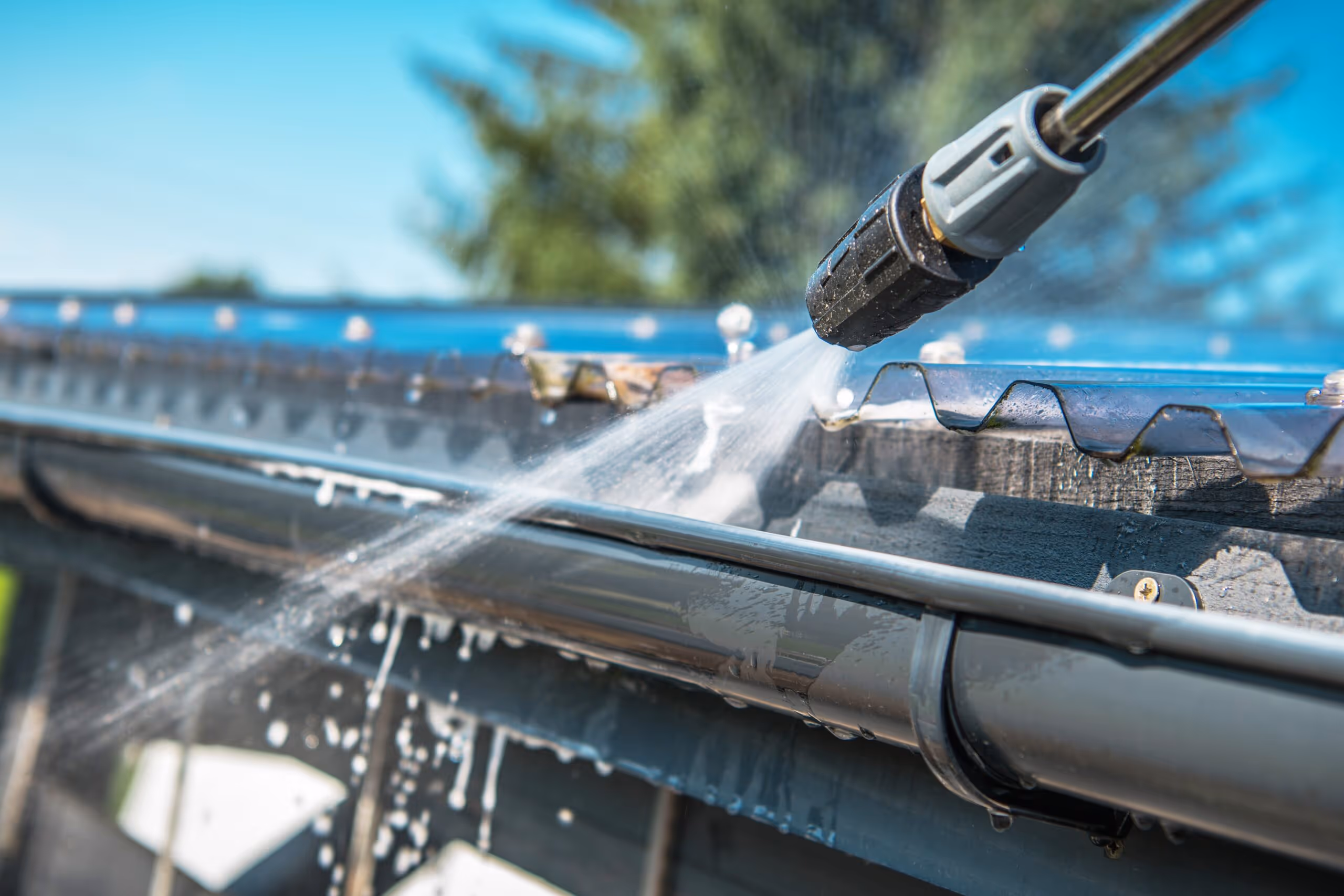 Pressure washer nozzle spraying water on a gutter to clean it on a sunny day.