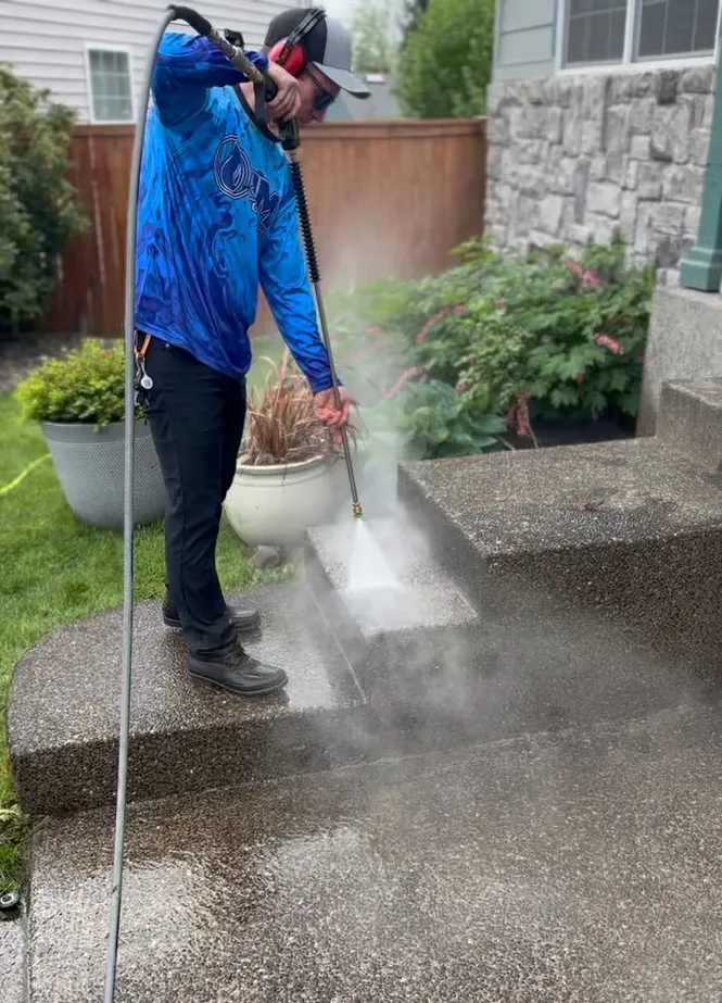 Man wearing a blue shirt and protective gear pressure washing concrete steps outdoors.