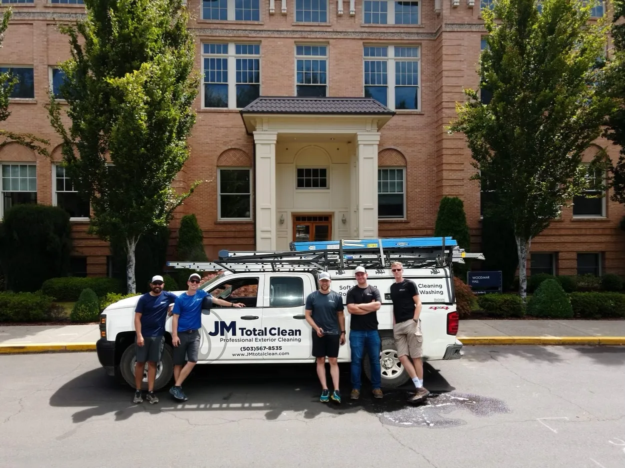 Five men standing and leaning against a white JM Total Clean truck parked in front of a brick building with green trees.