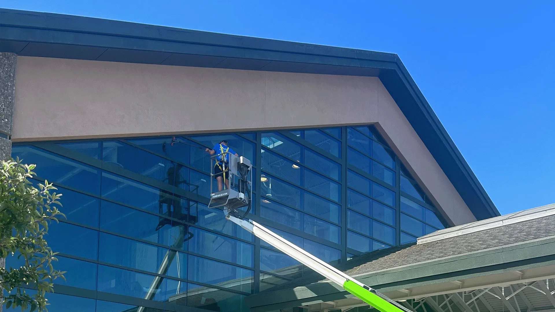 Worker wearing safety harness cleaning large glass windows on a building using an elevated platform lift under clear blue sky.