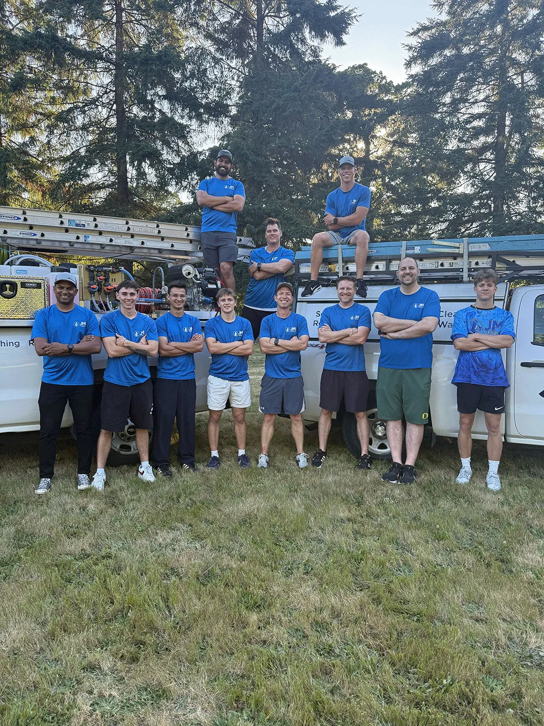 Group of eleven men in matching blue shirts posing with crossed arms in front of two white trucks loaded with ladders and equipment on a grassy area with tall trees in the background.