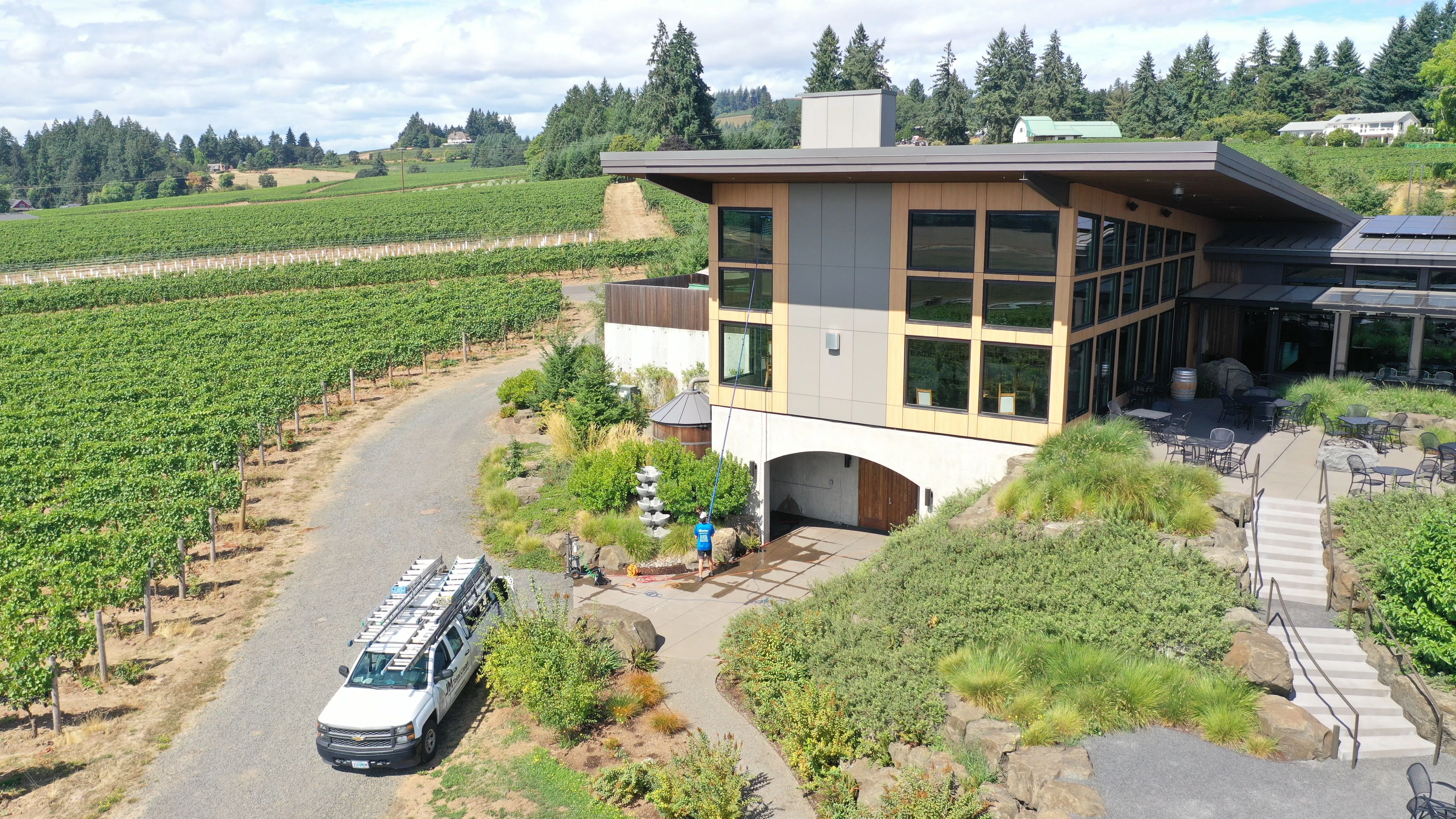Modern winery building next to vineyard rows with a person cleaning exterior and white utility truck parked nearby.