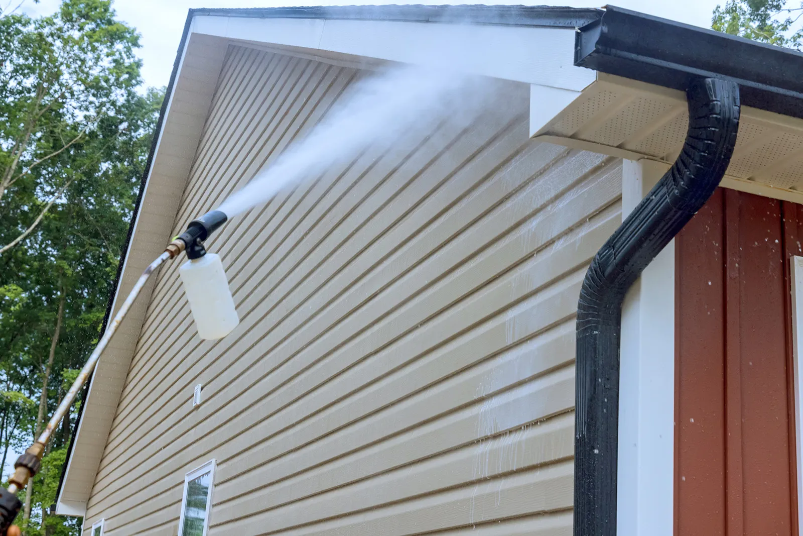 Pressure washer spraying water onto beige siding of a house near a black rain gutter.