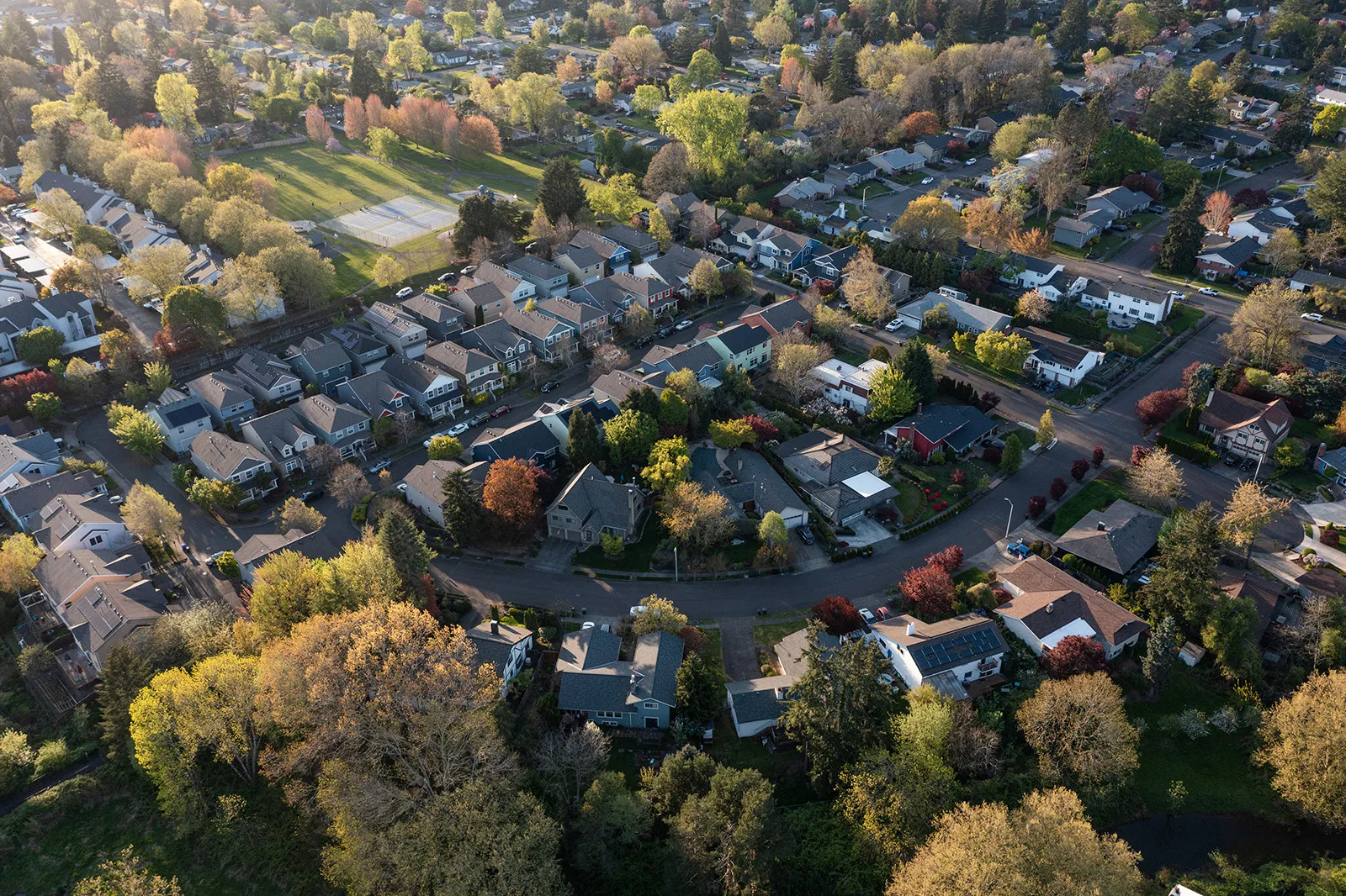 Aerial view of a suburban neighborhood with houses, trees in early autumn colors, and a park with tennis courts.