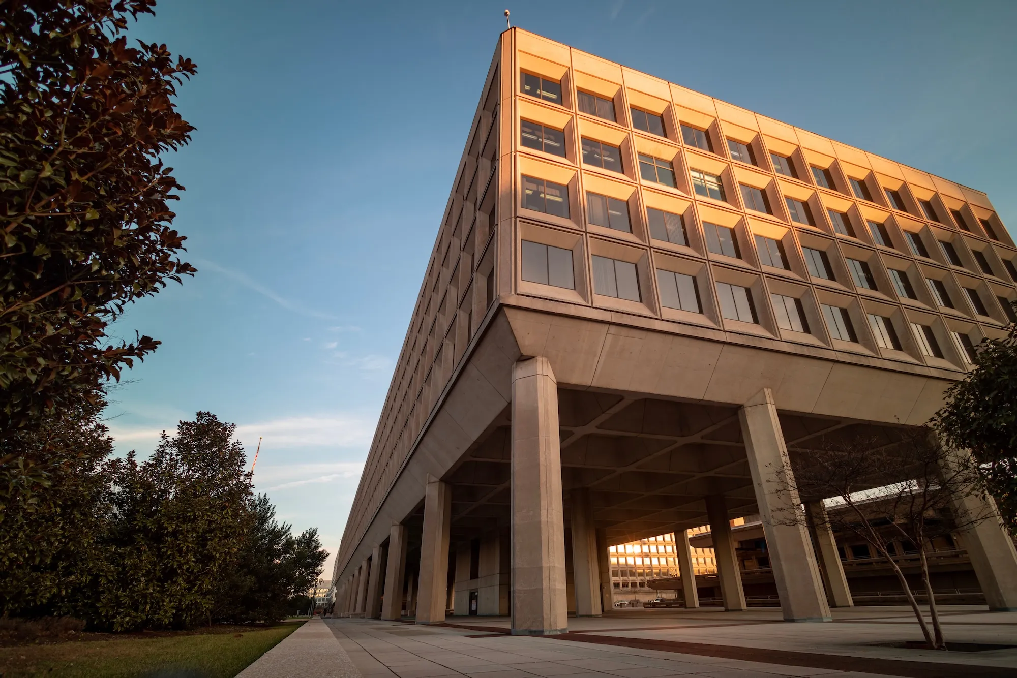 Brutalist concrete government building with grid-patterned windows lit by sunset, surrounded by trees and paved walkways.