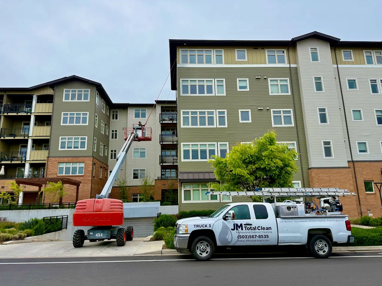 Professional exterior cleaning truck with equipment parked in front of a multi-story apartment building with a worker elevated on a lift cleaning windows.