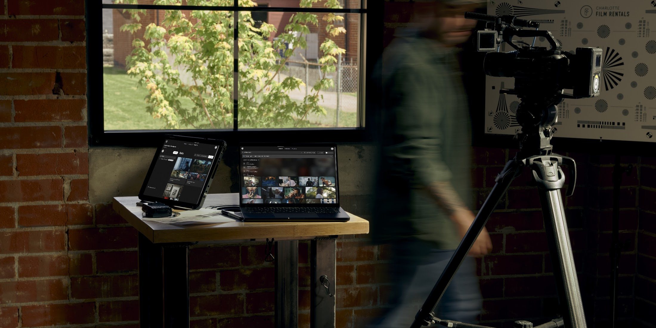 A filmmaker walking past a standing desk in a brick studio, with Frame Set open on an iPad and a laptop and a camera on a tripod nearby