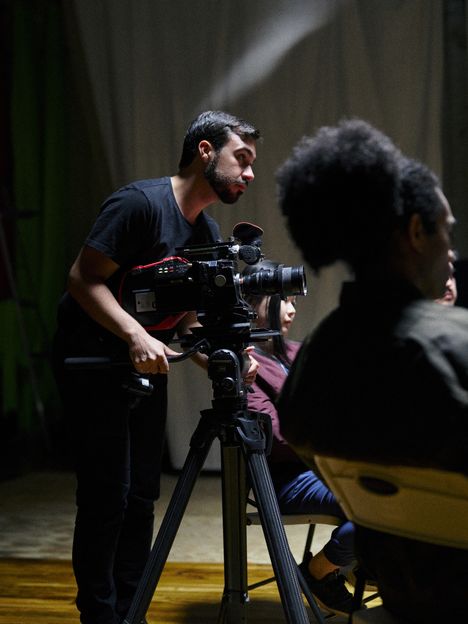 A camera operator frames a shot on a cinema camera while a crew member watches from behind