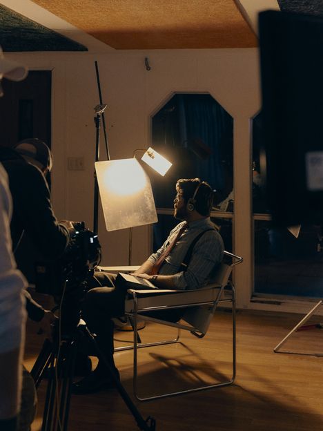 A performer sits in a chair under a single soft light while the crew works around them