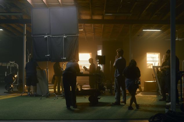 A crew works at the far end of a warehouse stage lit by practical lights