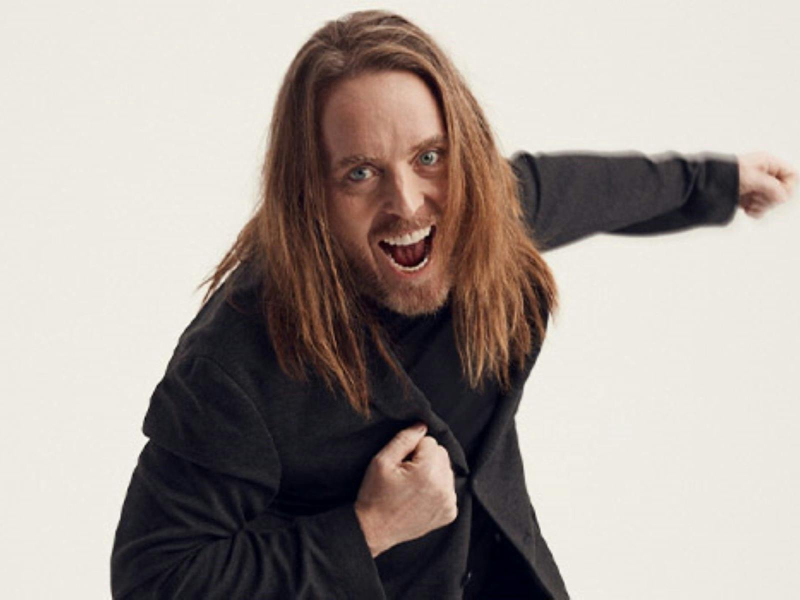 Photo of Tim in a black jacket and shirt with his hair down one arm out one  to his chest