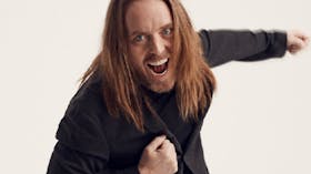 Photo of Tim in a black jacket and shirt with his hair down one arm out one  to his chest