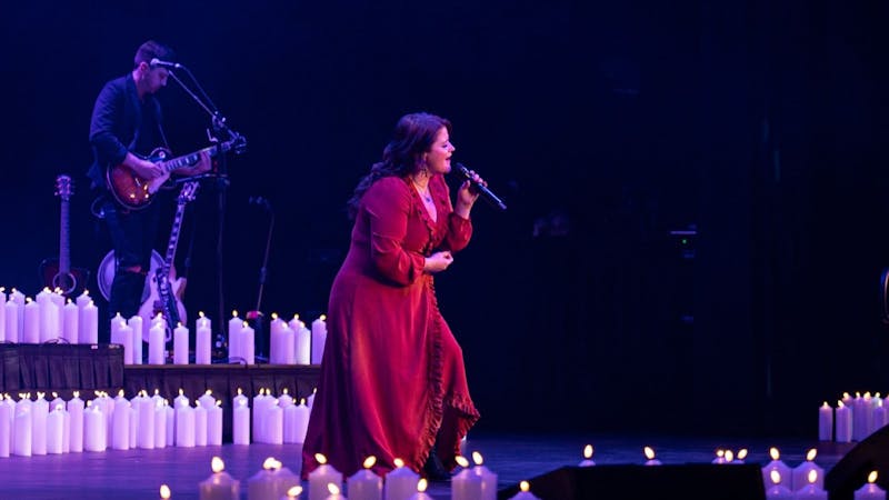 Photo of singer on stage wearing a red dress surrounded by candles holding the microphone on a stand