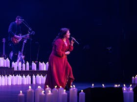 Photo of singer on stage wearing a red dress surrounded by candles holding the microphone on a stand