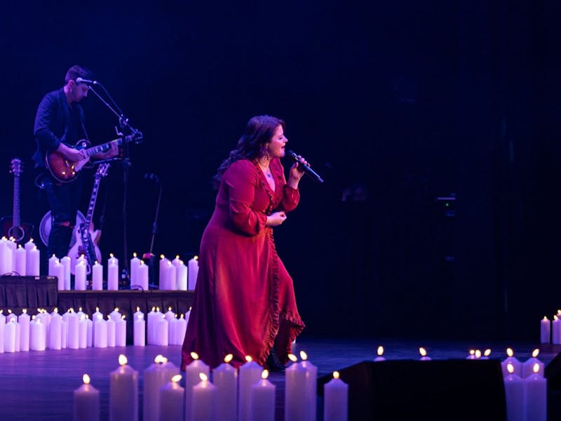 Photo of singer on stage wearing a red dress surrounded by candles holding the microphone on a stand