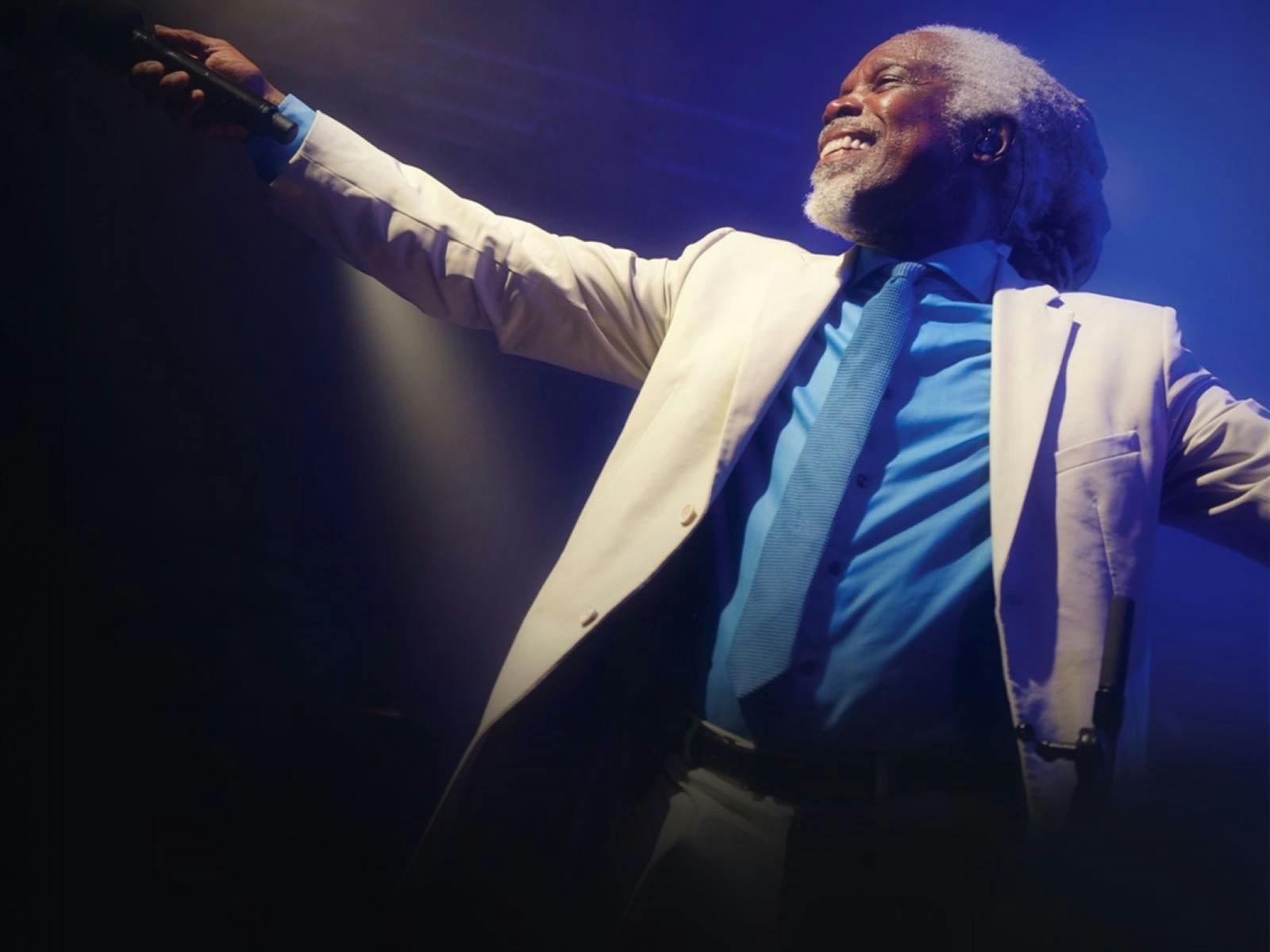 Photo of Billy Ocean in a white suit jacket and blue shirt and tie with his arm out