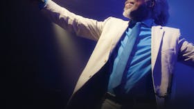 Photo of Billy Ocean in a white suit jacket and blue shirt and tie with his arm out