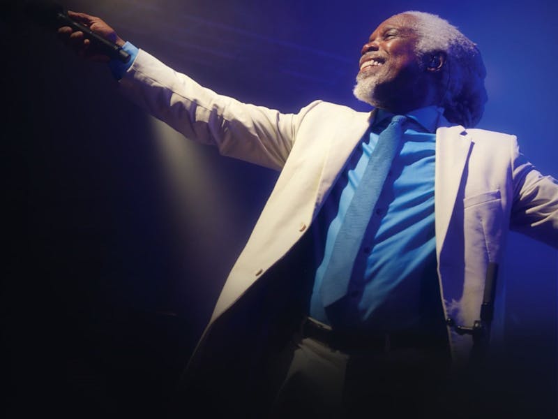 Photo of Billy Ocean in a white suit jacket and blue shirt and tie with his arm out