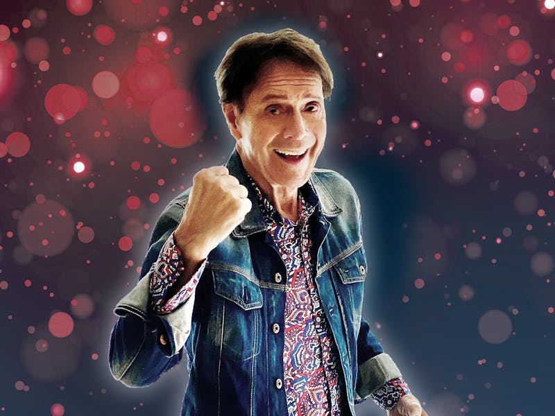 Photo of Cliff Richard smiling with his fist in a ball in front of a purple and pink bubble backdrop