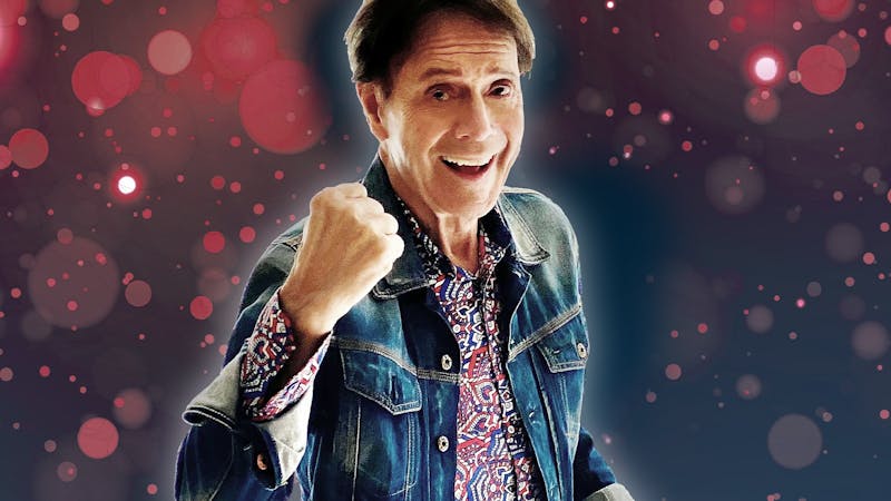 Photo of Cliff Richard smiling with his fist in a ball in front of a purple and pink bubble backdrop