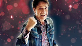 Photo of Cliff Richard smiling with his fist in a ball in front of a purple and pink bubble backdrop