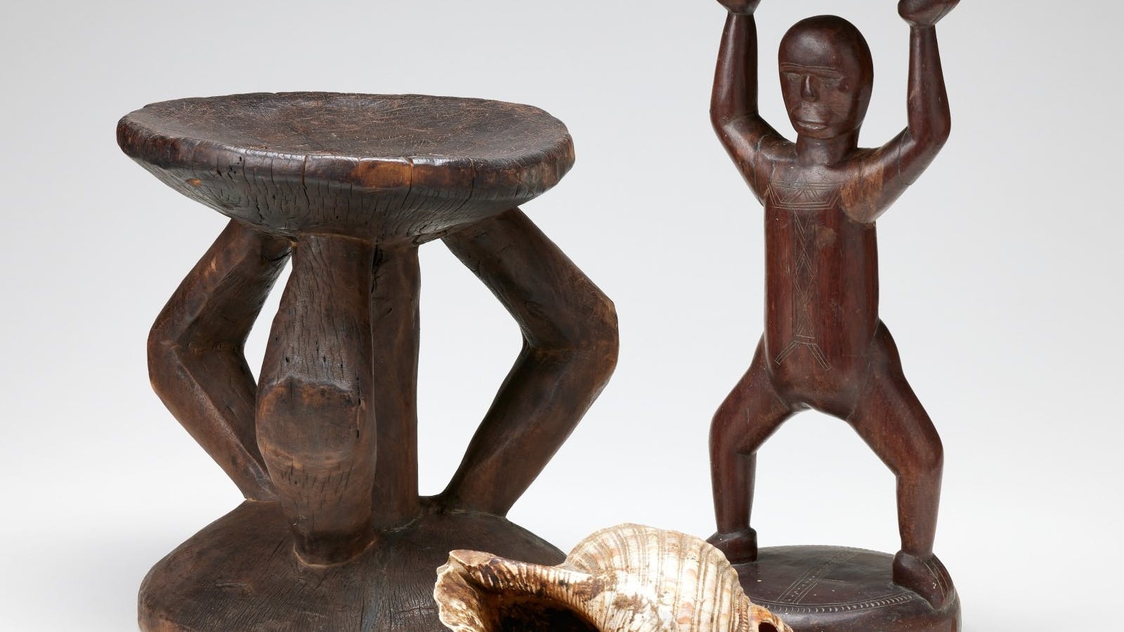 Two carved, wooden stools with one depicting the human form. A large shell with a small hole.