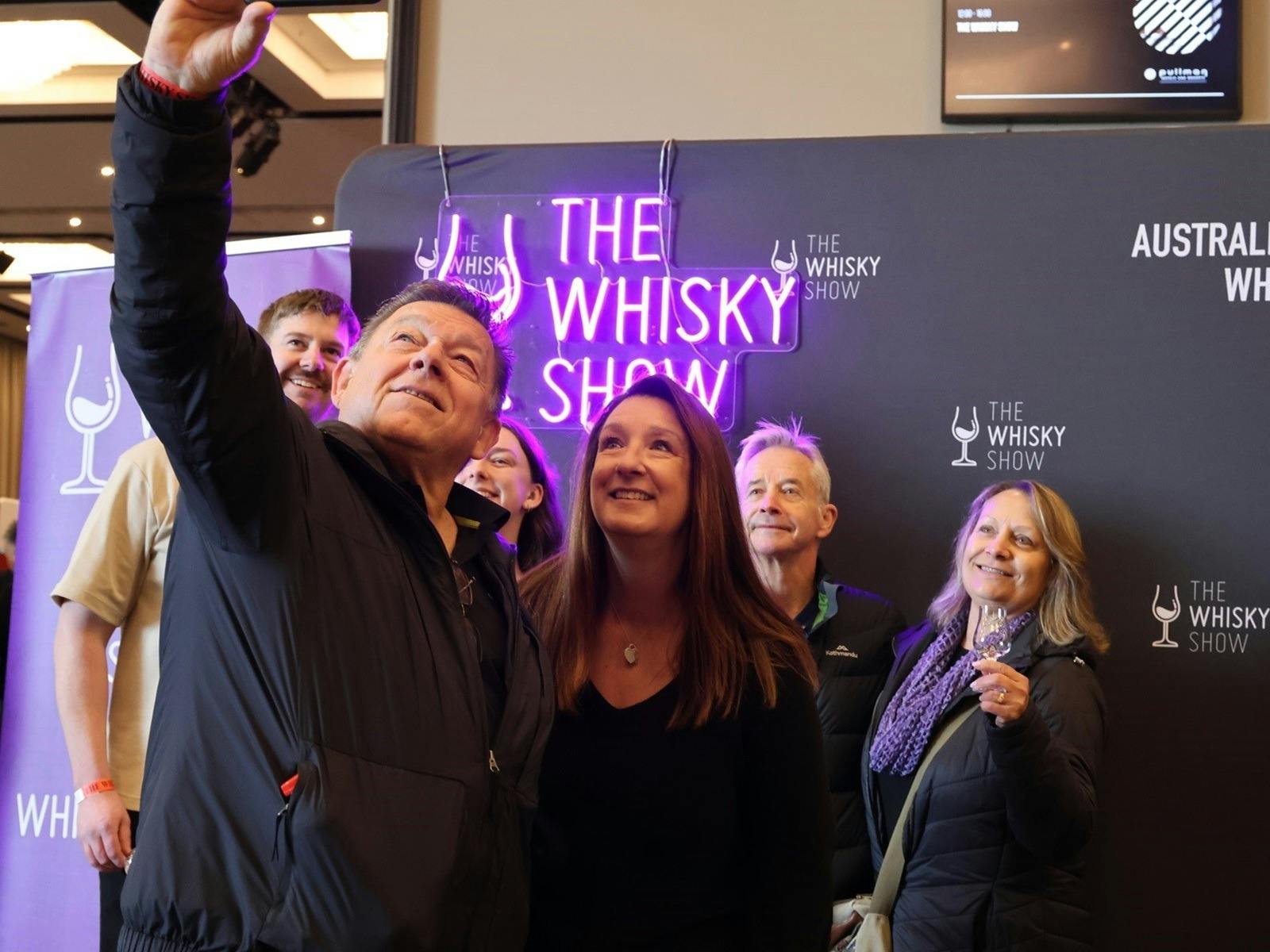 A group of friends gather in front of a Whisky Show banner and Neon Sign.