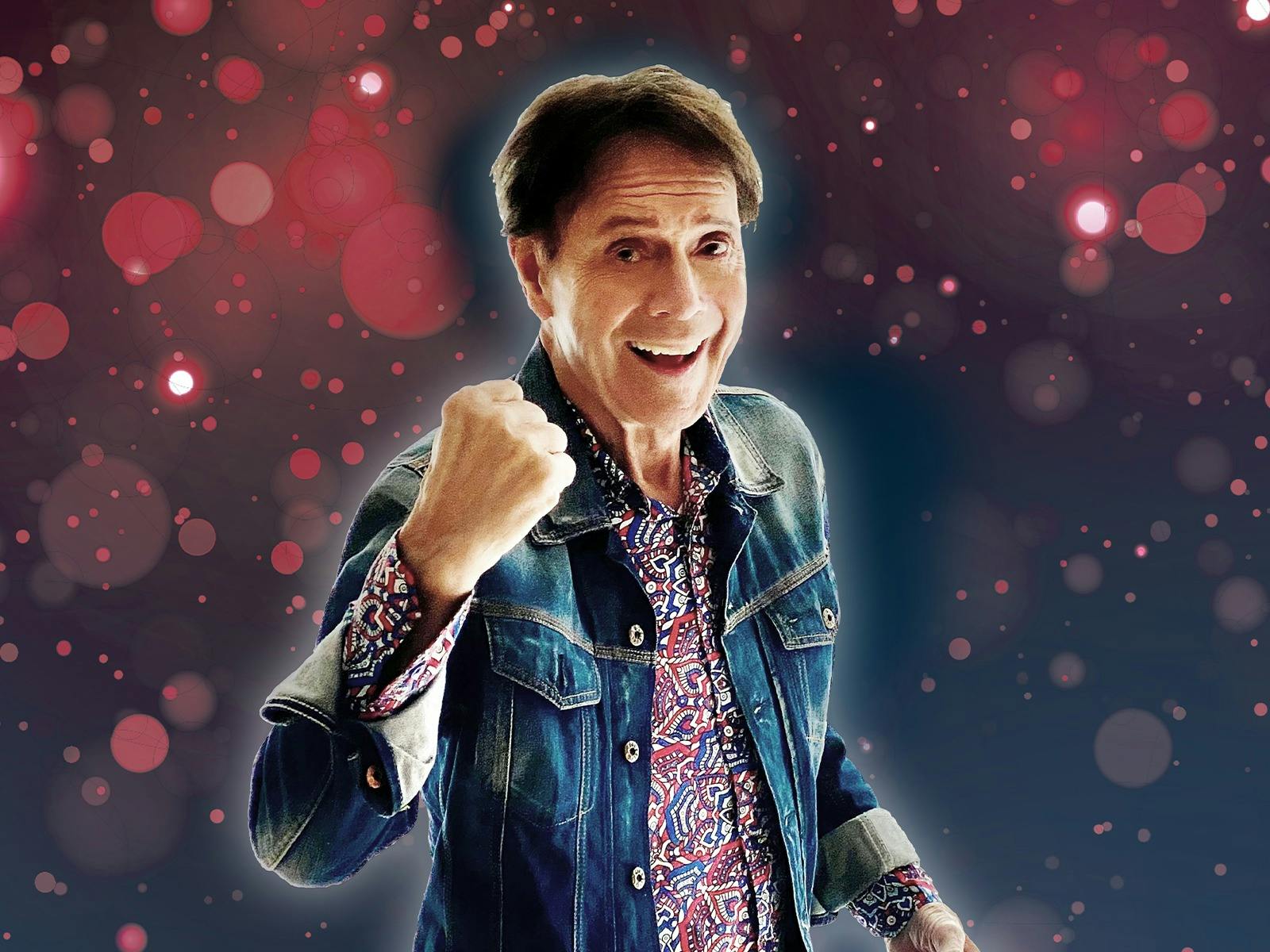 Photo of Cliff Richard smiling with his fist in a ball in front of a purple and pink bubble backdrop