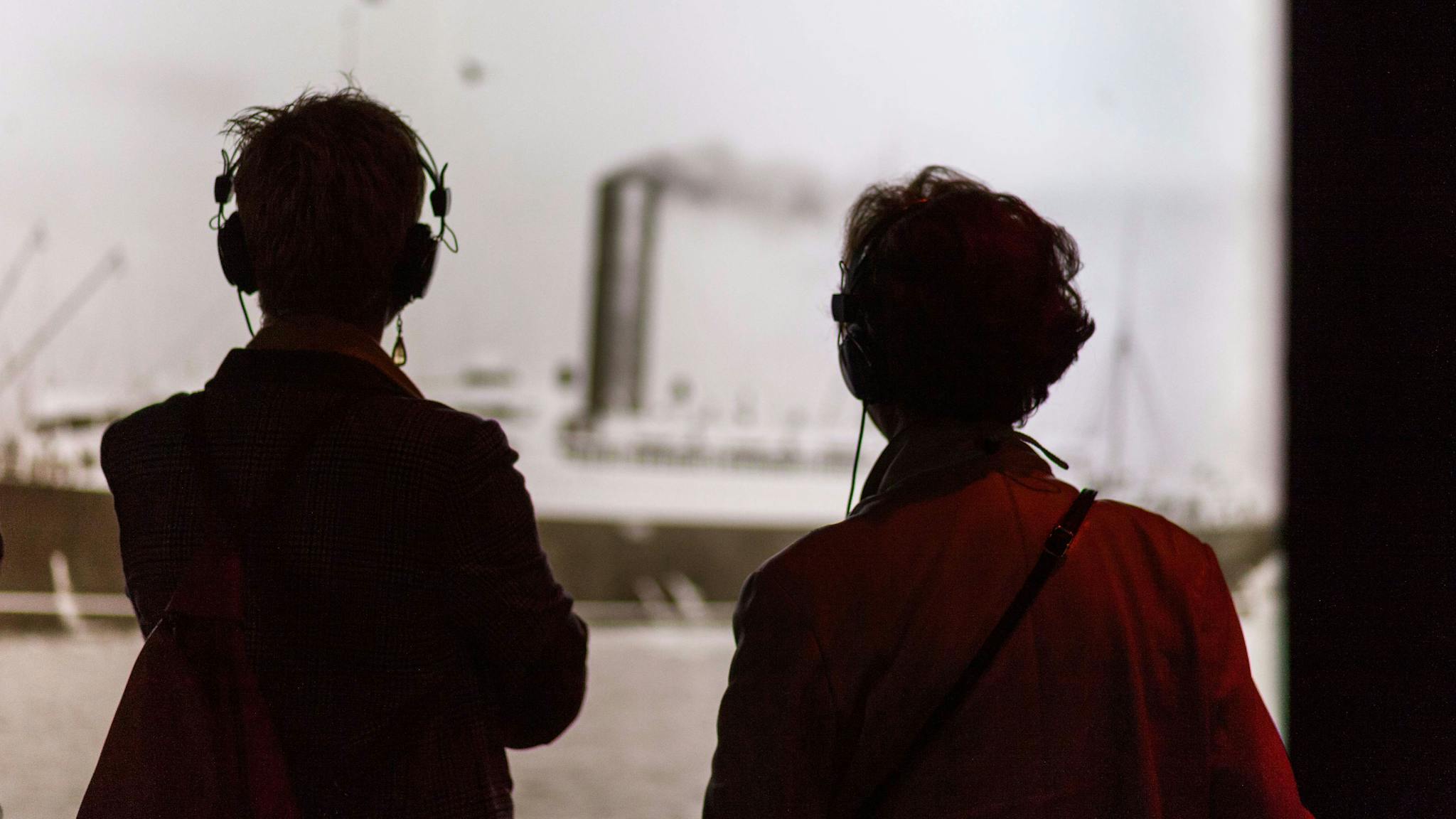 Two visitors onlook a photo while listening through guided headphones