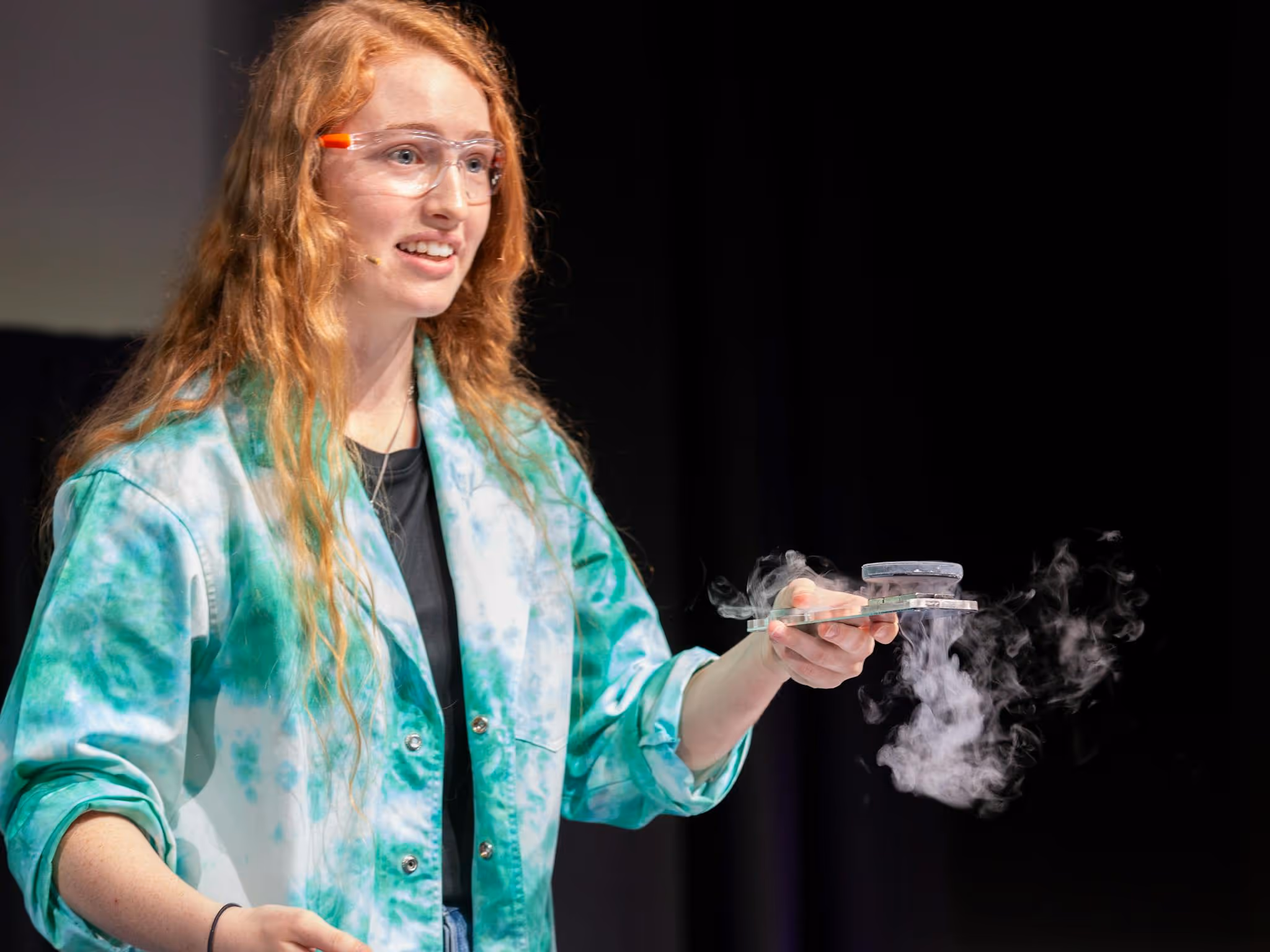 A woman with long red hair and a tye dye lab coat does a science experiment