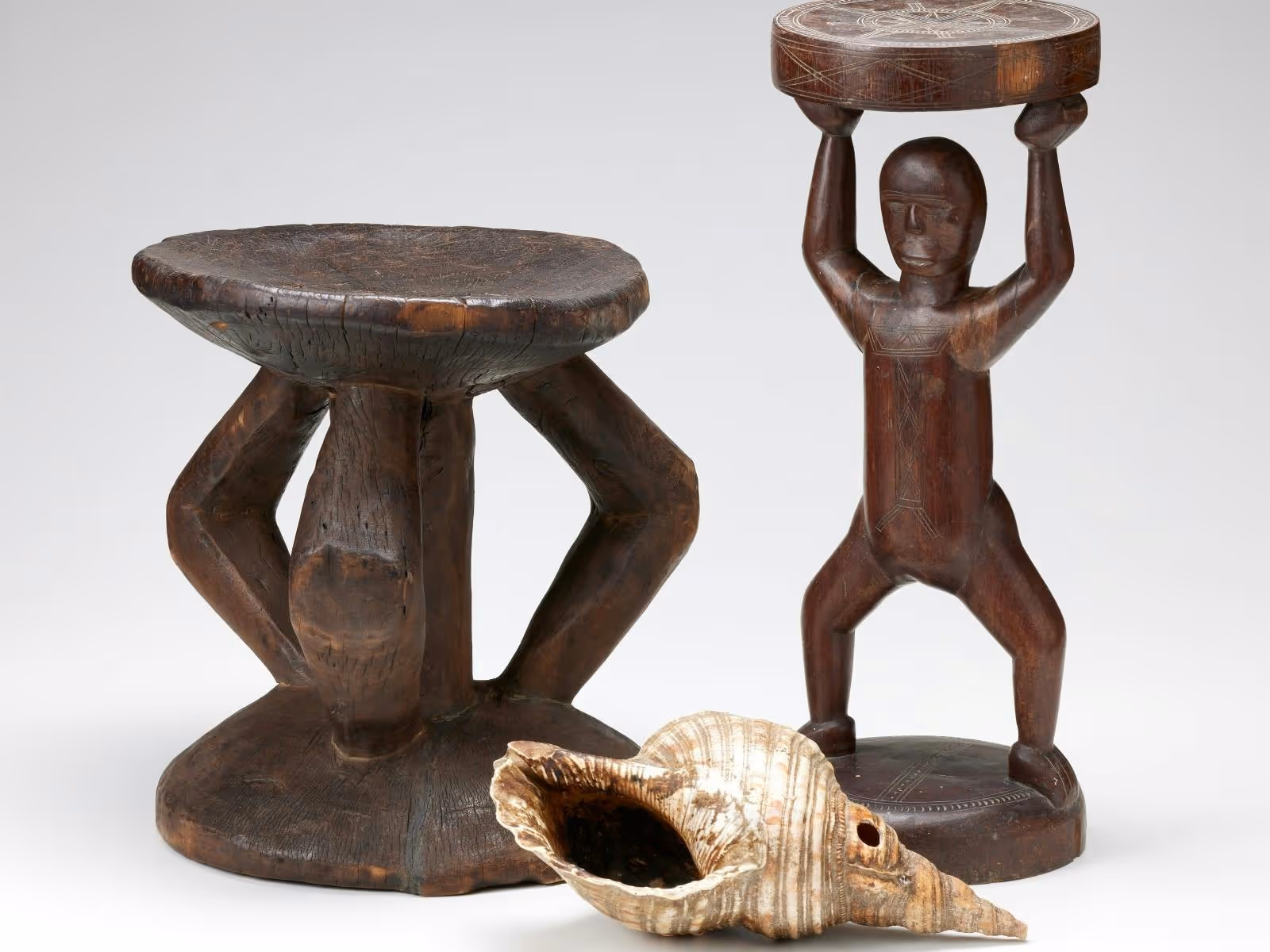 Two carved, wooden stools with one depicting the human form. A large shell with a small hole.