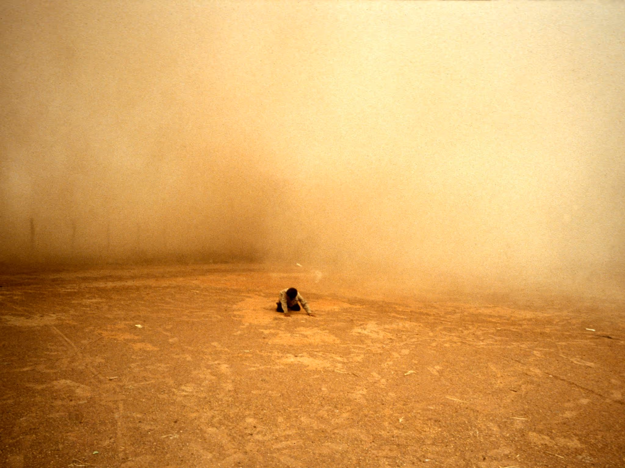 A person in the distance kneeling and looking downwards, surrounded by orage dust