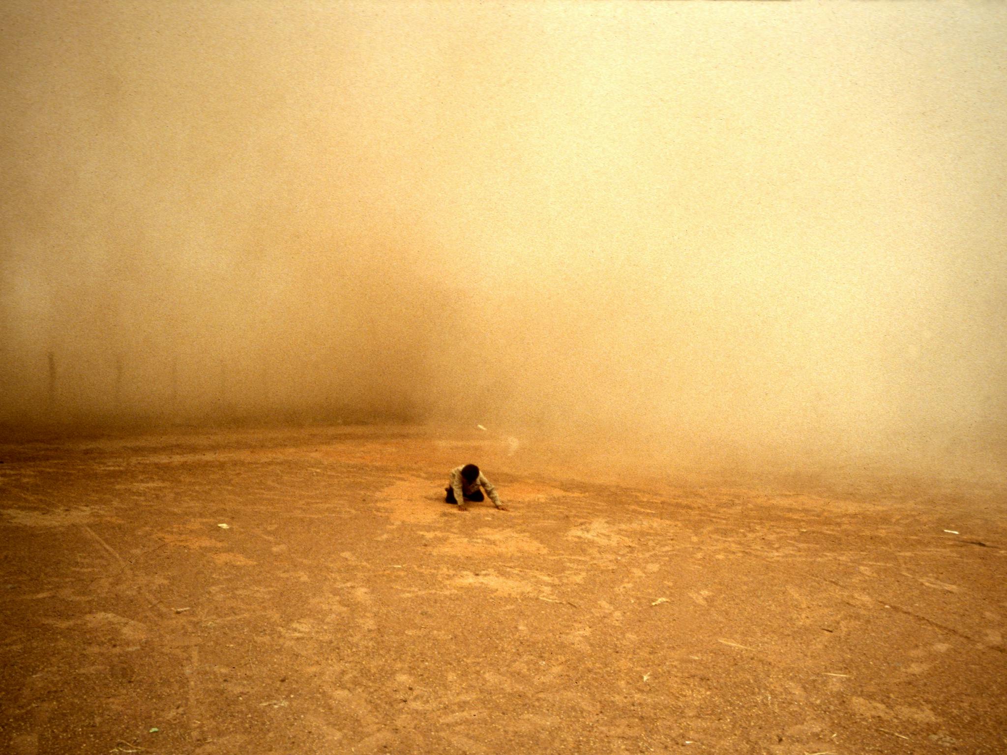 A person in the distance kneeling and looking downwards, surrounded by orage dust
