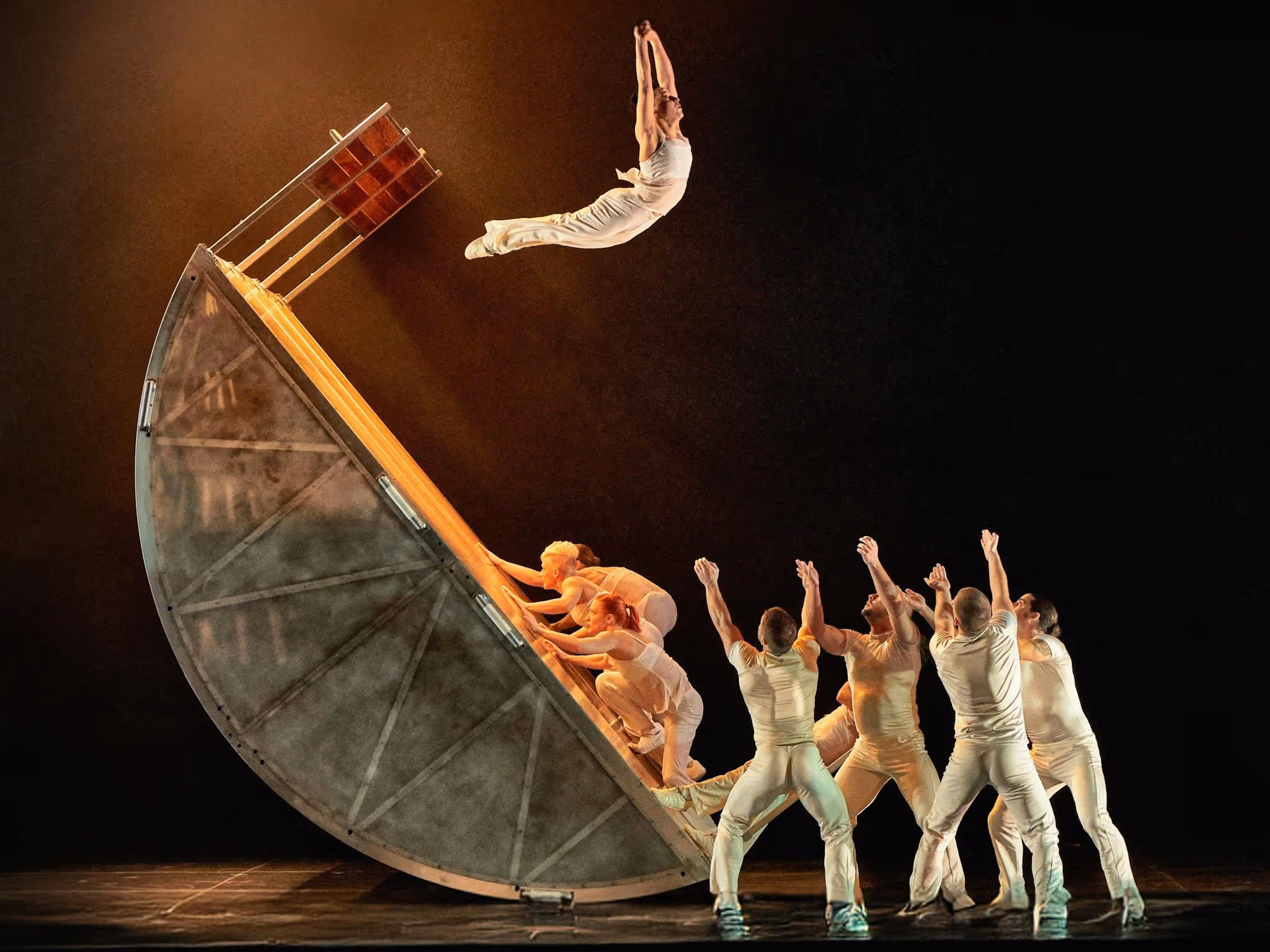 A performer flips off a semi-circular structure in mid air, while others stand below.