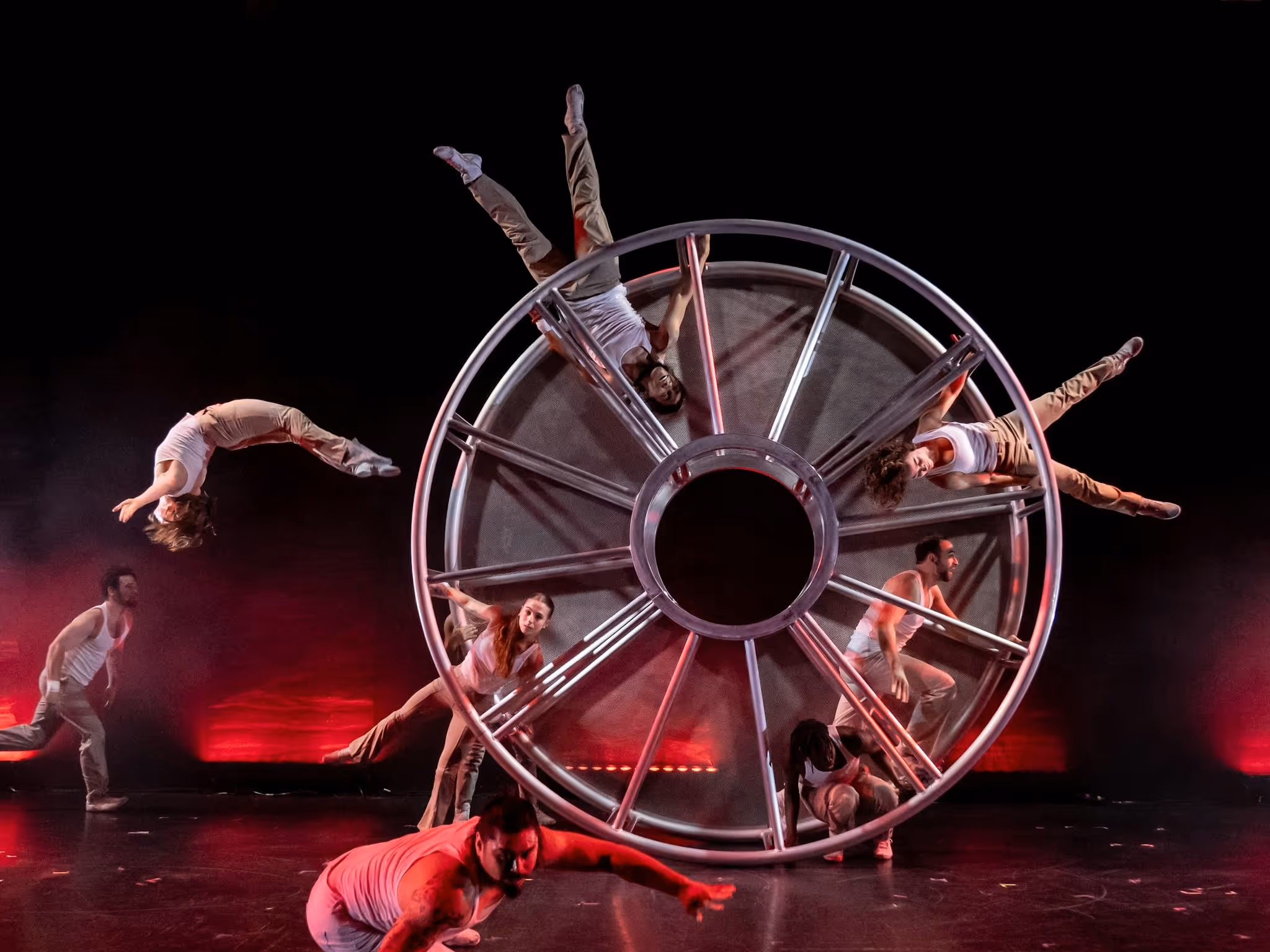 Circus performers are holding onto a wheel as it rotates. Red lighting