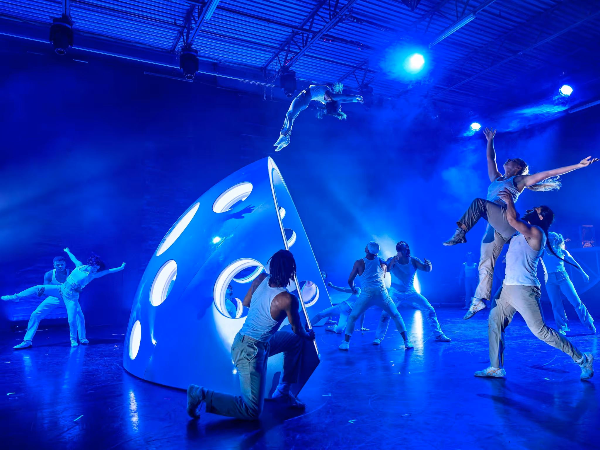 A group of performers are in motion around an architectural structure. The lights are blue.