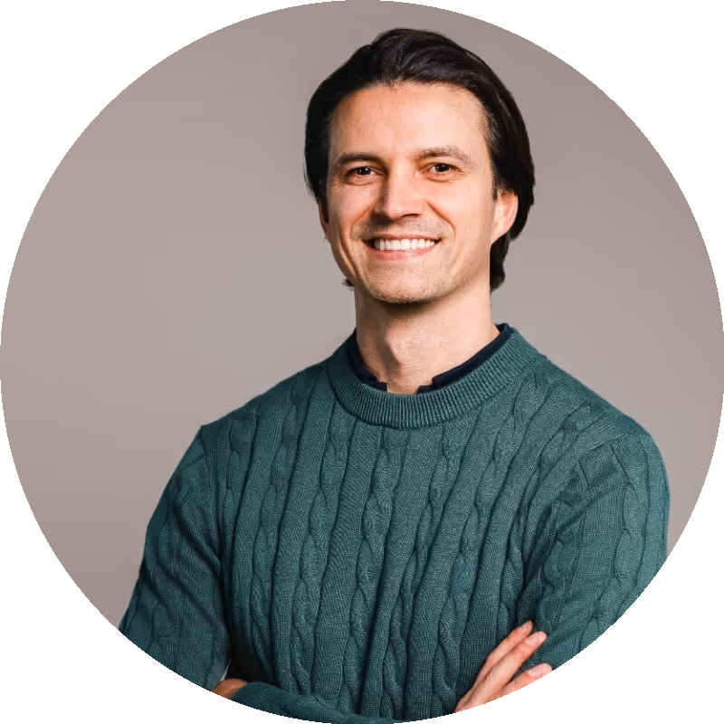 Hombre sonriente de cabello oscuro con un suéter de punto trenzado verde azulado, brazos cruzados, frente a un fondo neutro.