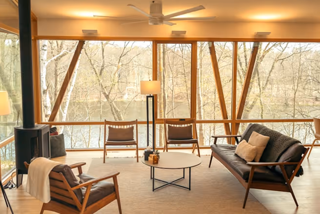 Modern living room with wooden furniture, large windows showing trees outside and a floor lamp.