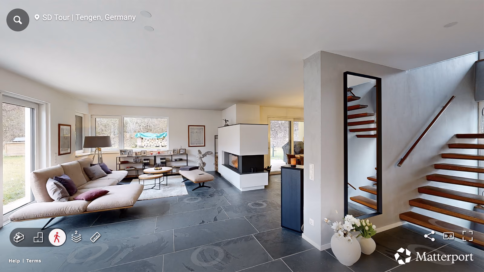 Modern living room with beige sofa, fireplace, wooden stairs, slate floor, and large windows.