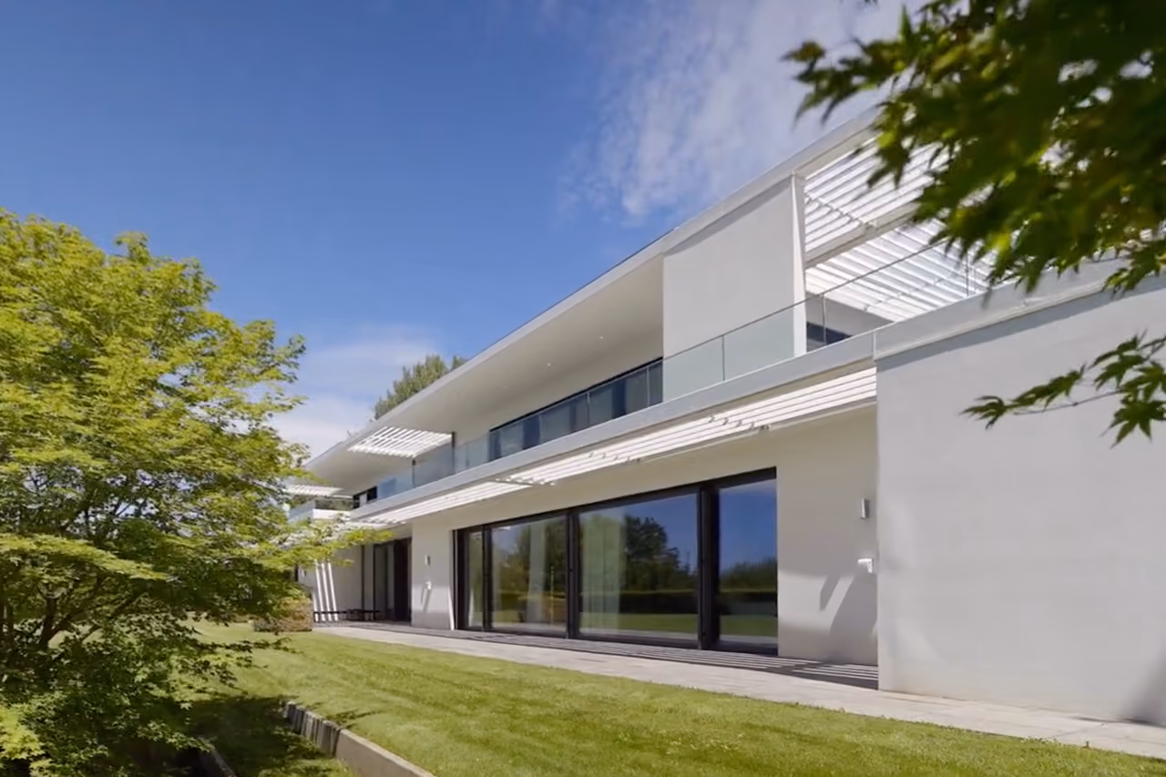 Modern two-story white house with large glass windows, balcony, and green lawn with trees.