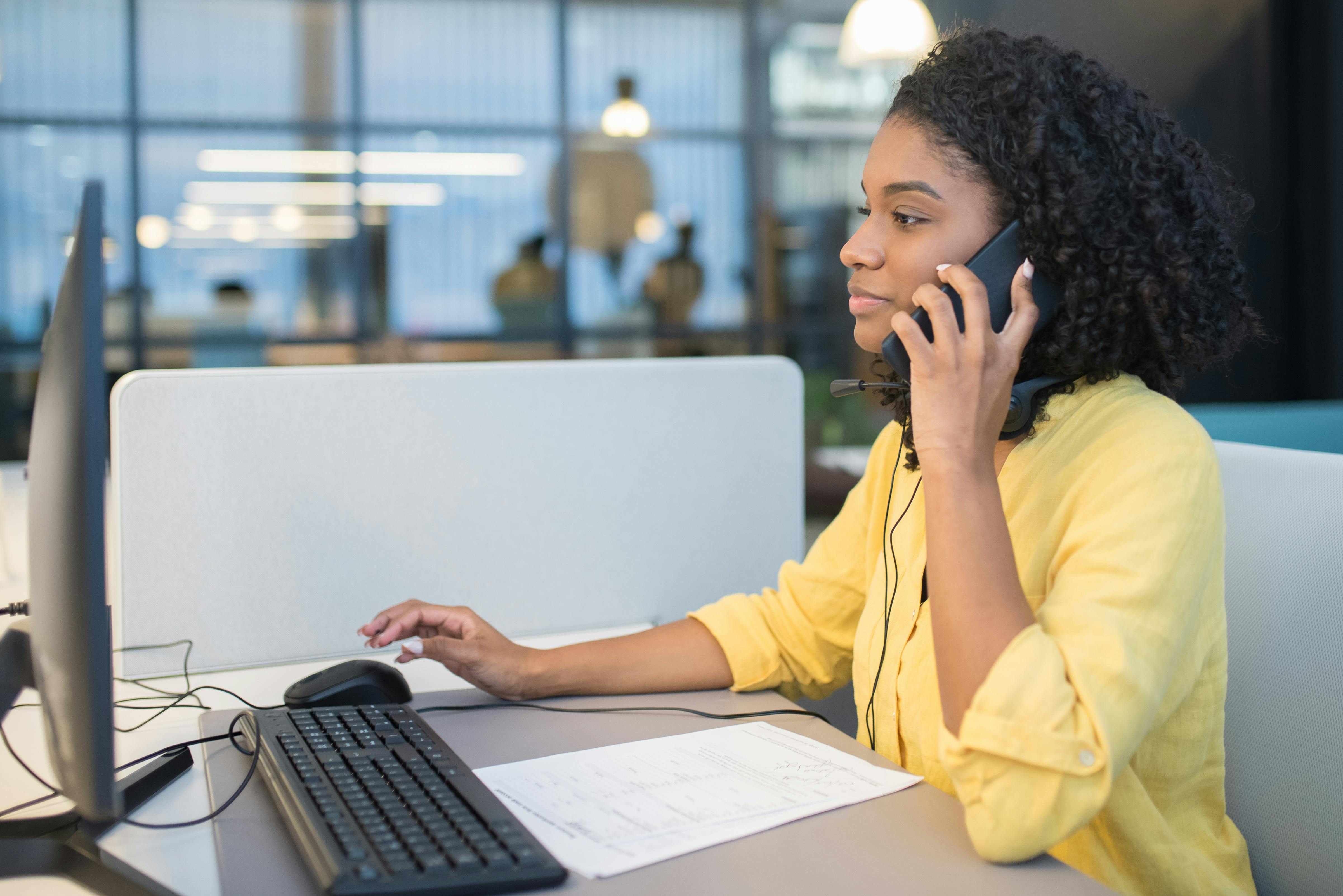 Customer support agent speaking on the phone while using a desktop computer. Represents how attackers target third-party support staff through multiple channels - including voice, email, and chat - to compromise enterprise access.