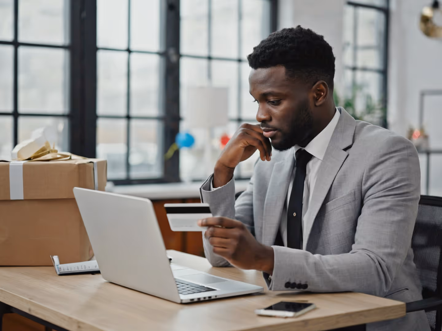 Man in an office holding a credit card while checking his laptop, with shipping boxes and subtle holiday decorations in the background, illustrating how workplace online shopping can increase exposure to seasonal scams and fraudulent payment requests. 
