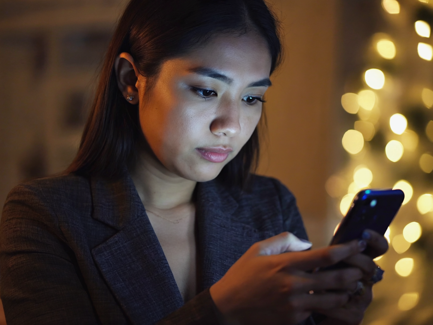 Close up of a woman in a business blazer looking intently at her smartphone, with soft holiday lights blurred in the background, illustrating how employees can receive suspicious texts or scam messages during the holiday season while working.