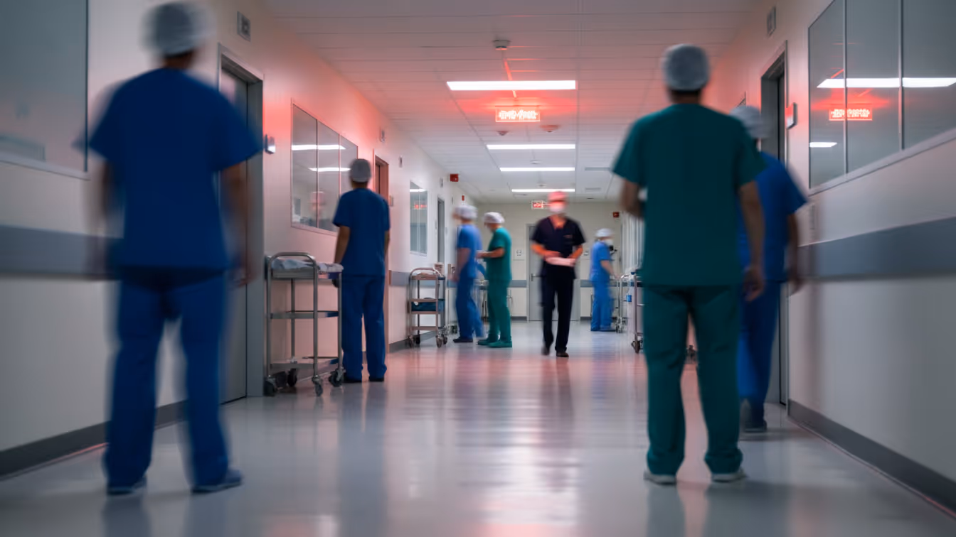 Blurred hospital corridor with multiple healthcare staff in scrubs walking and working, viewed from the center of the hallway. Red emergency lights glow overhead, conveying a busy, high-pressure clinical environment with active hospital operations. 