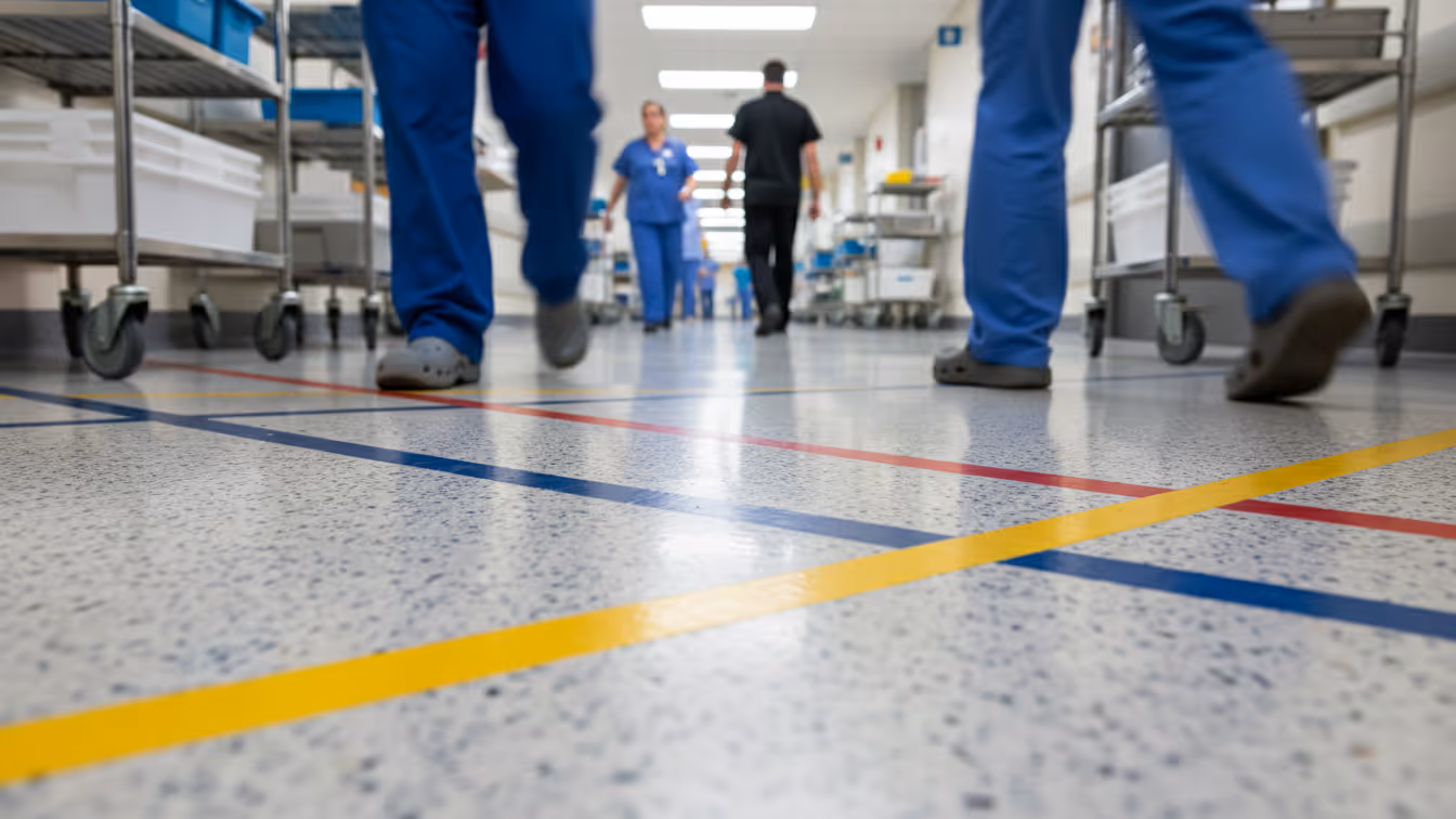 Low-angle view of a hospital corridor floor with colored guidance lines, as healthcare staff walk through the hallway in the background, showing active movement and day-to-day hospital operations. 