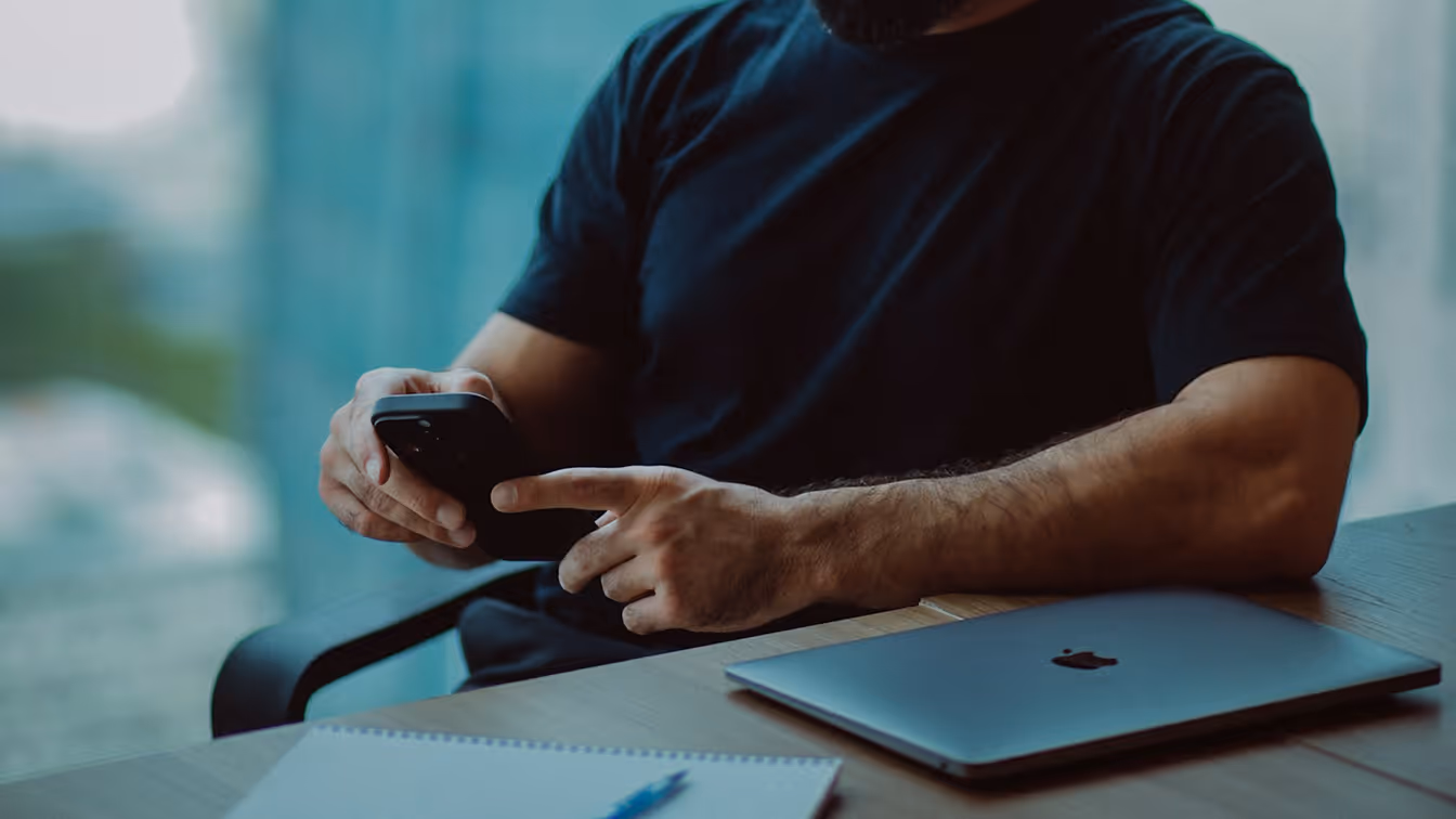Person sitting at a desk using a smartphone next to a laptop, representing interaction with digital systems and workflows.